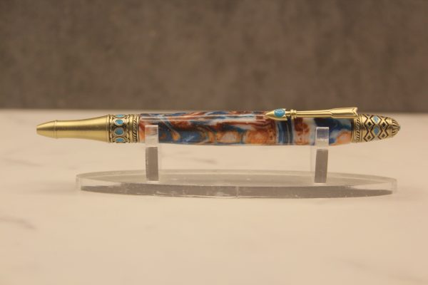 resin pen with gold-colored patterned hardware laying horizontally on a plastic pen stand, writing tip facing left. The hardware has southwestern-adjacent zigzags, diamonds, circles, and scallop patterns molded into it, and the pen clip is in the shape of a stylized feather arrow, and all of the hardware is accented with turquoise colored resin set into the design. The resin body is one piece with swirled colors including turquoise, white, and orangey-gold