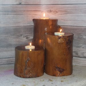 three 4 inch thick orange-brown sealed wooden cylinders with preserved wood imperfections with a lit tealight inlaid in each candle holder. The one in the back is 8 inches tall, the shortest of the two in front of it (the one on the left) is 4 inches tall, and the one on the right is 6 inches tall