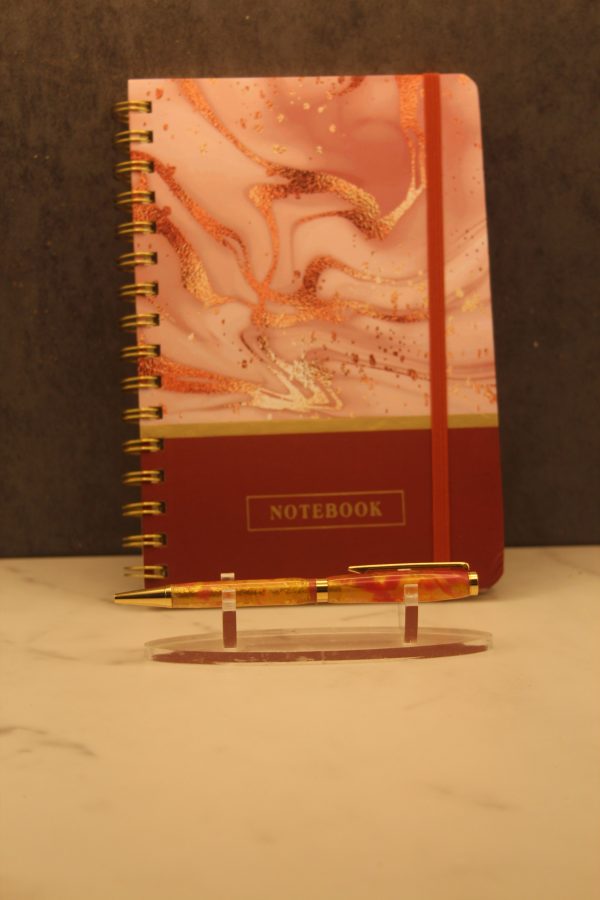 Fire themed notebook and pen set; gold wire-o bound notebook with a pink and gold marbling on the top three-quarters of the cover, and a red stripe on the bottom quarter, divided by a thin gold stripe. The red stripe features the word "notebook" typed in gold capital letters framed by a thin gold rectangle, and the notebook also comes with a red elastic band. In front of the notebook on a plastic stand is a handmade pen with gold attachments and two gold and red marbled segments.