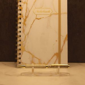 Air themed notebook and pen set; white notebook with gold colored marbling, gold wire-o type binding, and a gold elastic band. Centered towards the top of the cover is the word "Notebook" in gold typed script surrounded by a gold decorative frame. In front of the notebook on a plastic stand is a handmade pen with gold attachments and two white and tan marbled segments.