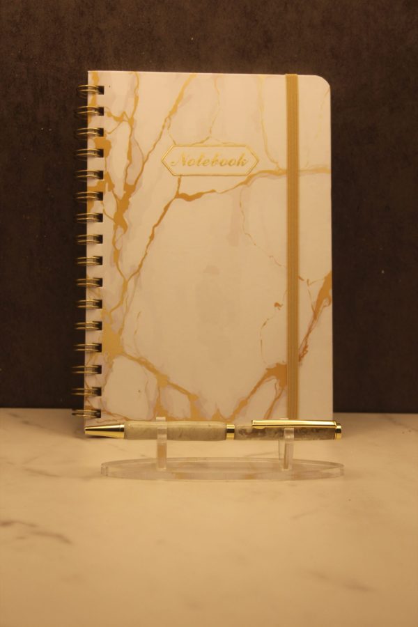 Air themed notebook and pen set; white notebook with gold colored marbling, gold wire-o type binding, and a gold elastic band. Centered towards the top of the cover is the word "Notebook" in gold typed script surrounded by a gold decorative frame. In front of the notebook on a plastic stand is a handmade pen with gold attachments and two white and tan marbled segments.