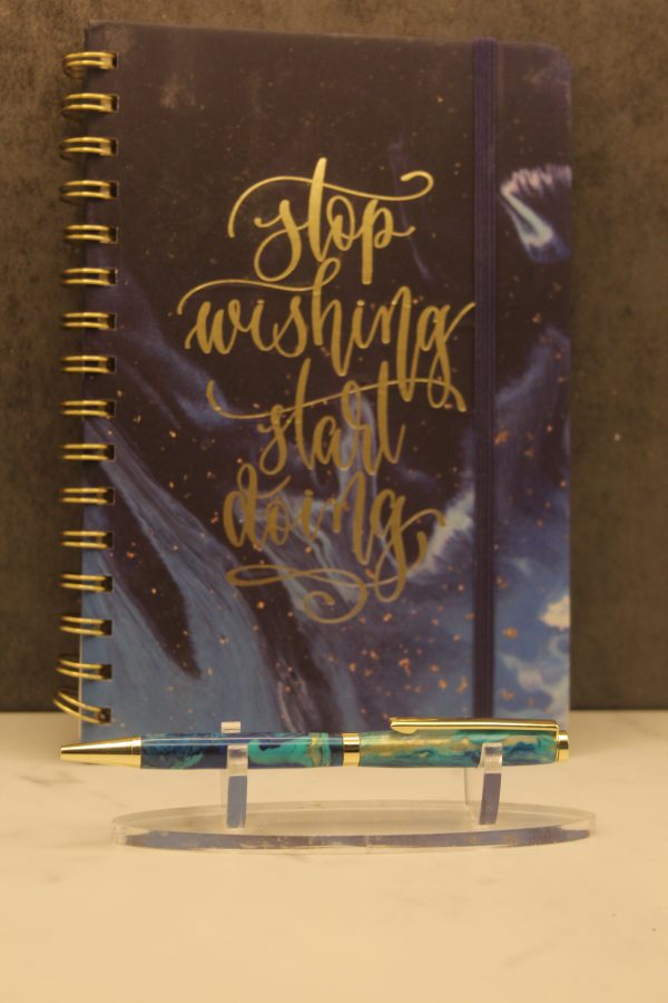 Water themed notebook and pen set; gold wire-o bound notebook with a navy blue and light blue marbling on the cover. Written on the cover in gold modern calligraphy script are the words "stop wishing start doing". The notebook also comes with a navy blue elastic band. In front of the notebook on a plastic stand is a handmade pen with gold attachments and two navy blue, turquoise, and gold marbled segments.