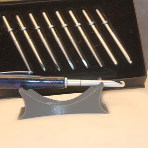 Silver metal crochet hook with a pen shaped handle including a matching pen clip, made of a purple, blue, and black marbled resin dubbed the "amethyst" colorway. Behind it is a black, velvet lined box with 8 other silver colored metal crochet hook heads in varying sizes