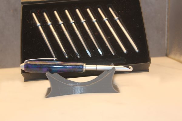 Silver metal crochet hook with a pen shaped handle including a matching pen clip, made of a purple, blue, and black marbled resin dubbed the "amethyst" colorway. Behind it is a black, velvet lined box with 8 other silver colored metal crochet hook heads in varying sizes