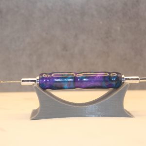 2 in 1 seam ripper and needle threader. This purple, blue, and black marbled resin handle is referred to as the "Amethyst" colorway, and has silver hardware. About the size of a pen, on one end is a seam ripper, and on the other end is a needle threader, which is a small wire loop. Both ends can be removed from the resin handle and reversed inside of itself for storage