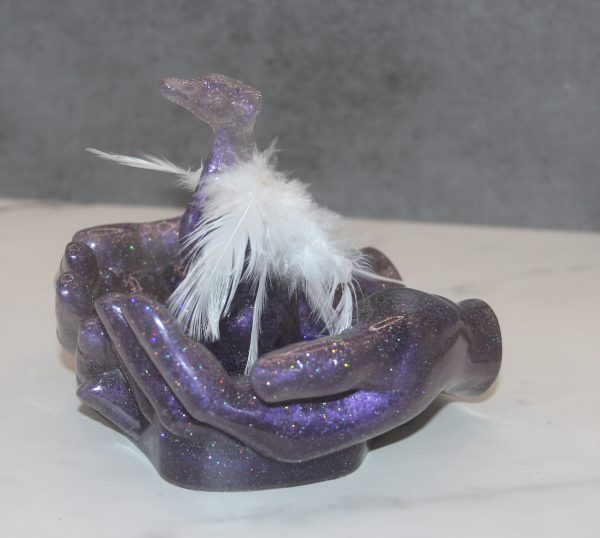 In His Hands is a sculpture of a purple sparkly whippet with feather angel wings being held by matching hands