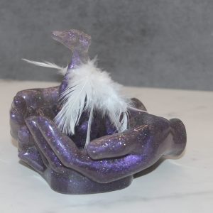 In His Hands is a sculpture of a purple sparkly whippet with feather angel wings being held by matching hands