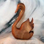Hand carved wooden swan sculpture with graceful curved neck and layered wing feathers, carved from rich honey-brown reclaimed wood, displayed against a marble backdrop