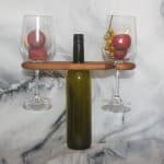 Italian olivewood wine caddy in use holding a green wine bottle and two stemmed glasses balanced by their bowls on marble surface